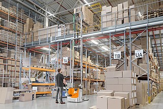 Person operating an automated pallet stacker in a large warehouse with tall shelves filled with boxes and labeled sections