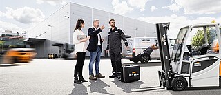 Three people are standing in front of a STILL warehouse. Surrounding them are several STILL forklift trucks.