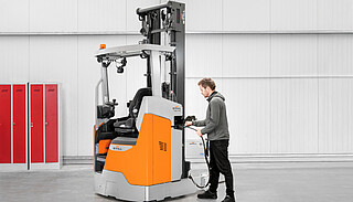 A man connects a charging cable to an orange and gray forklift in a brightly lit hall.