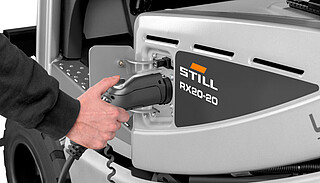 Close-up of a hand inserting a charging plug into the side charging socket of a silver forklift (model STILL RX20-20).