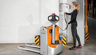 A woman in a warehouse connects the charging cable of an orange and silver electric pallet jack to a wall socket